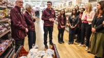 Group of supermarket staff and executives, including Andy Burnham and Sadiq Khan, standing in a bakery aisle.