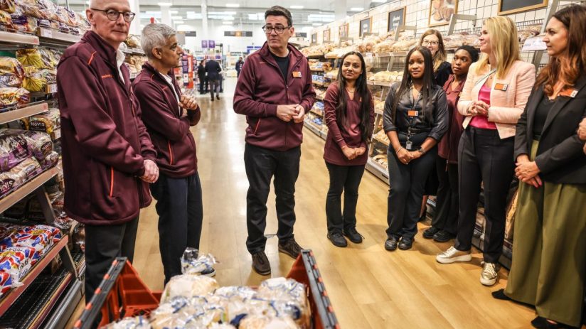 Group of supermarket staff and executives, including Andy Burnham and Sadiq Khan, standing in a bakery aisle.