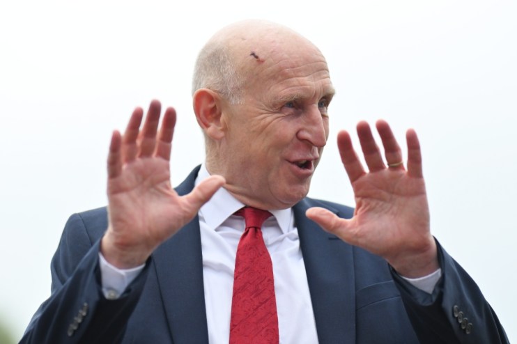 Bald man in suit and red tie gesturing with open hands, small scab visible on his forehead