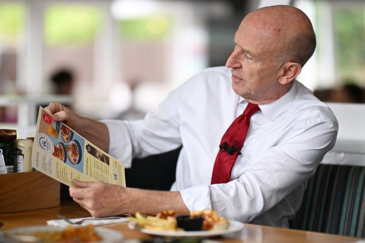 Bald man in white shirt and red tie reading a restaurant menu with a lapel microphone attached