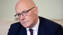 Bald man in glasses and dark suit with a purple tie and Scottish flag pin, looking to the left.