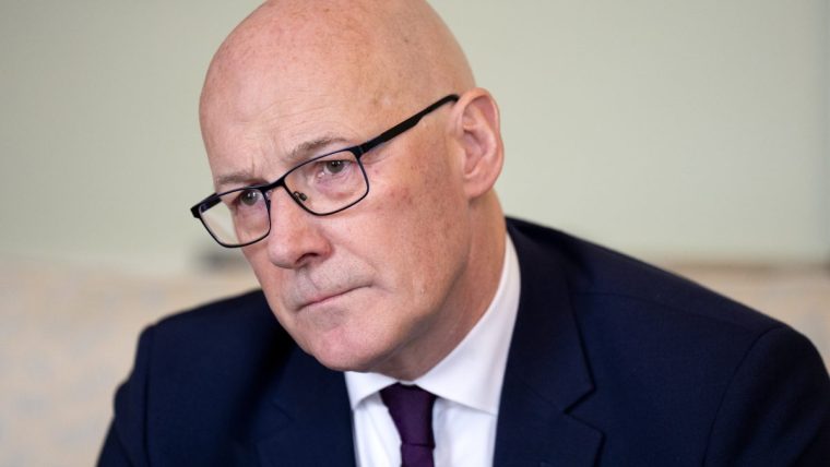 Bald man in glasses and dark suit with a purple tie and Scottish flag pin, looking to the left.
