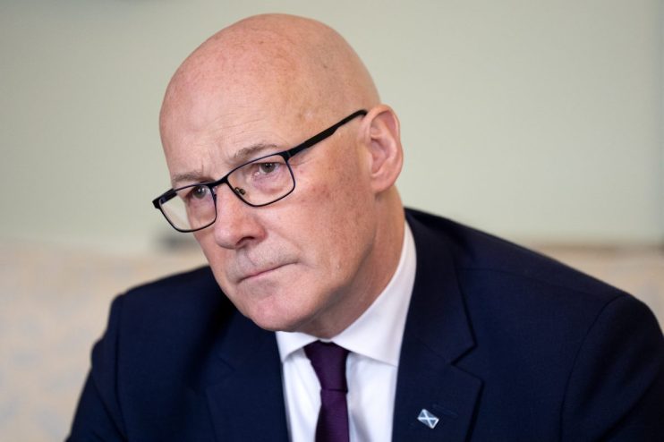 Bald man in glasses and dark suit with a purple tie and Scottish flag pin, looking to the left.