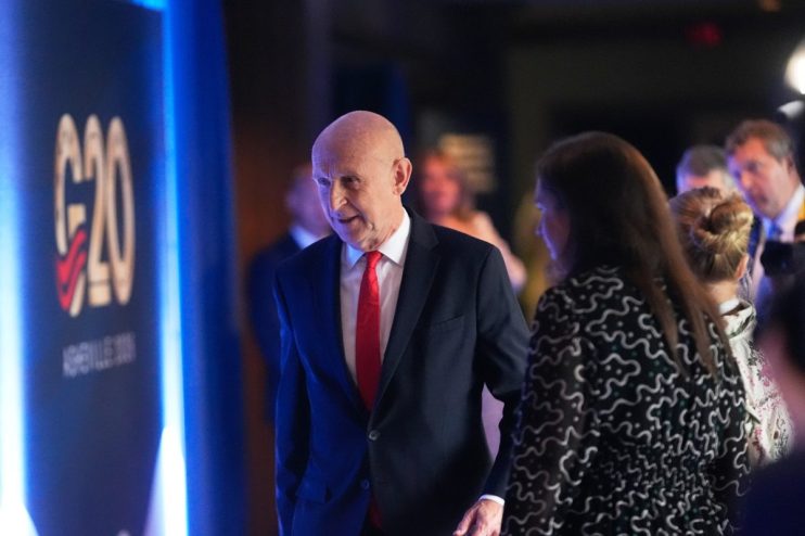 Bald man in a dark suit and red tie walking past a G20 sign with other attendees in background.