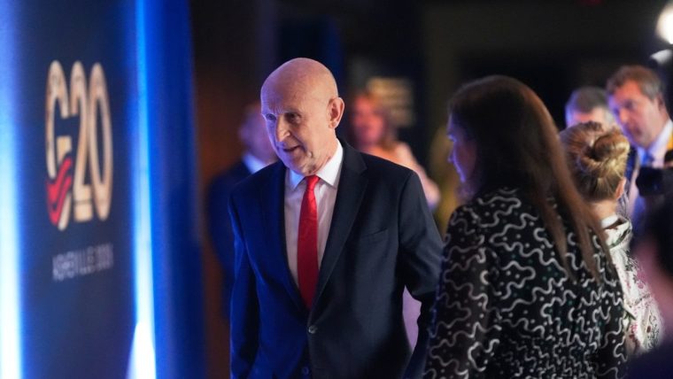 Bald man in a dark suit and red tie walking past a G20 sign with other attendees in background.