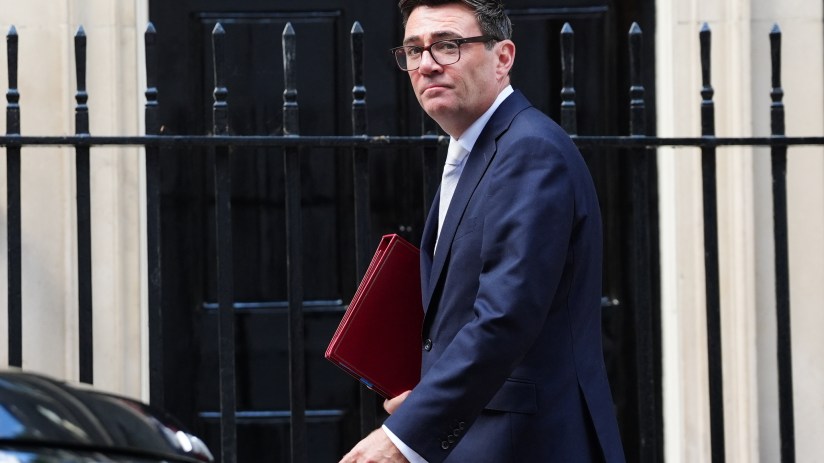 Andy Burnham, Mayor of Greater Manchester, in a suit, holding a red folder, walking past railings