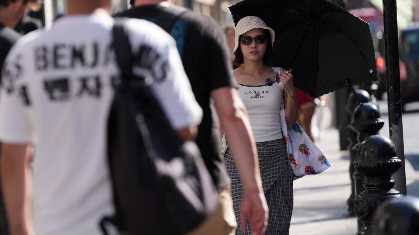 A woman in sunglasses and a hat holding a black umbrella on a sunny city street, with blurred people in the foreground.