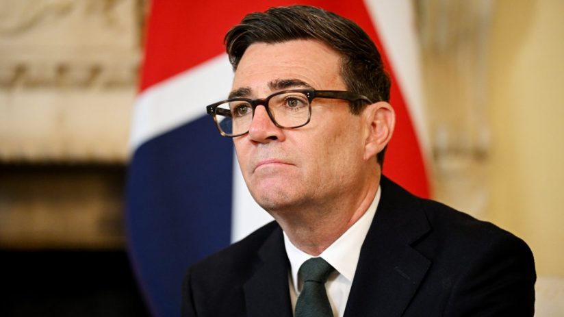 Andy Burnham, Mayor of Greater Manchester, in a suit and tie with a Union Jack flag in the background.