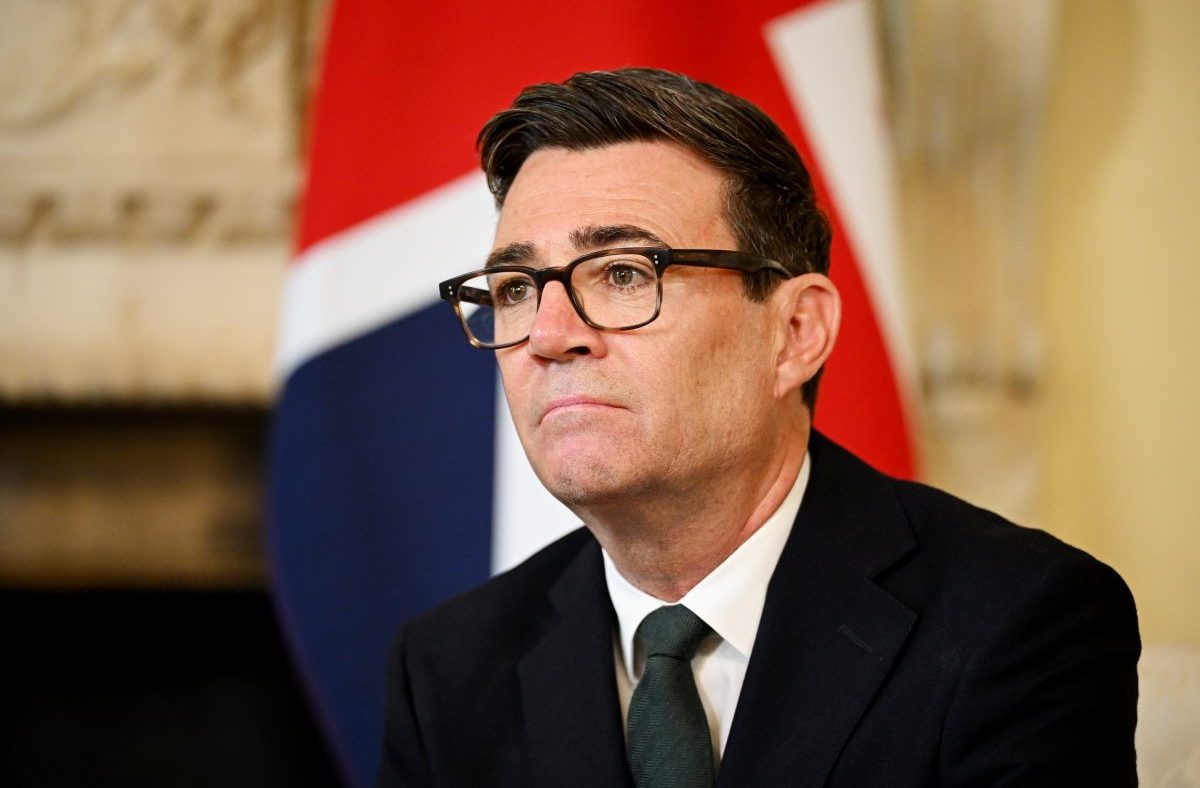 Andy Burnham, Mayor of Greater Manchester, in a suit and tie with a Union Jack flag in the background.