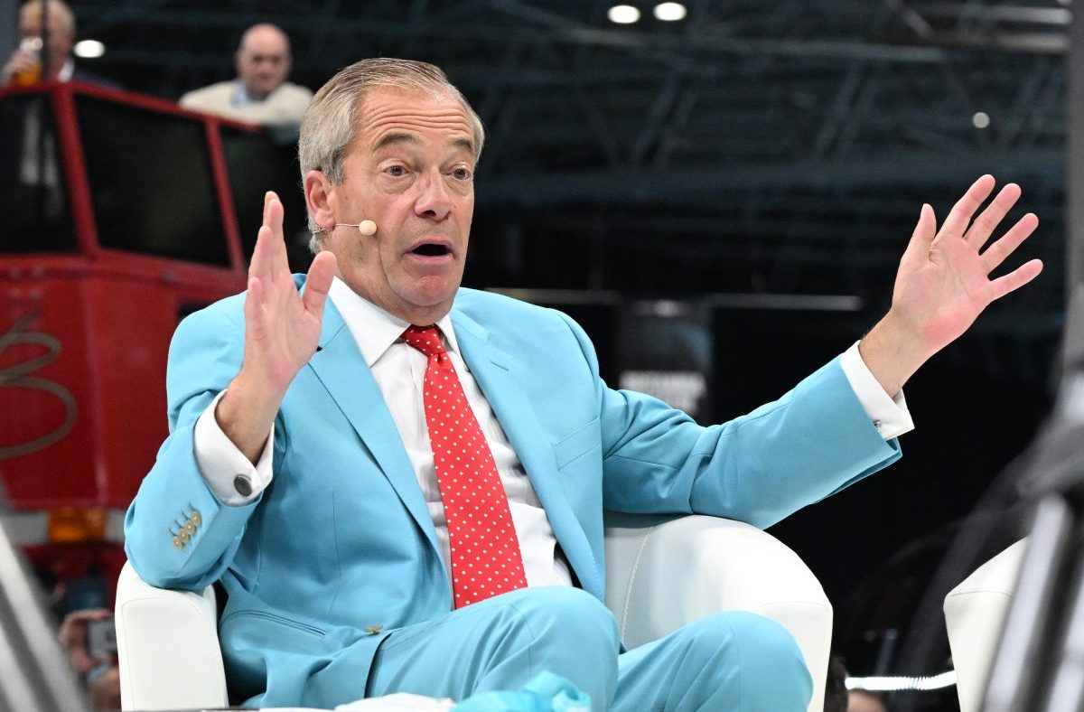 Nigel Farage in a light blue suit and red polka dot tie, speaking with hands raised, at a public event.