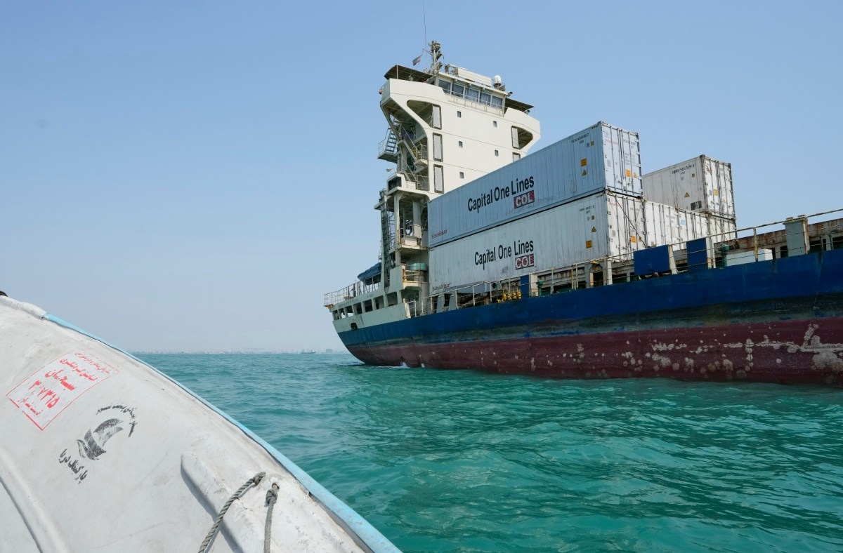 Large cargo ship, Capital One Lines containers visible, on bright blue water, with a small boat in foreground.