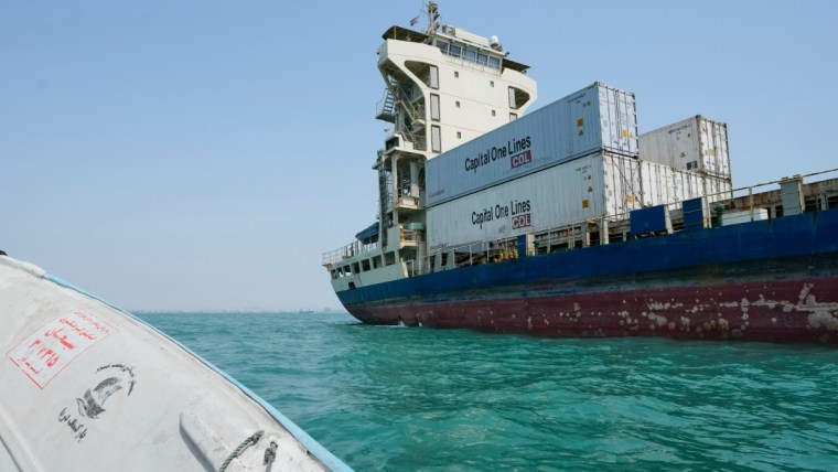 Large cargo ship, Capital One Lines containers visible, on bright blue water, with a small boat in foreground.
