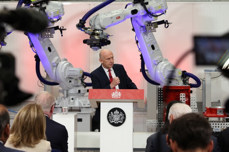 A bald man in a suit speaking at a podium with the Royal Coat of Arms, flanked by two ABB industrial robots.