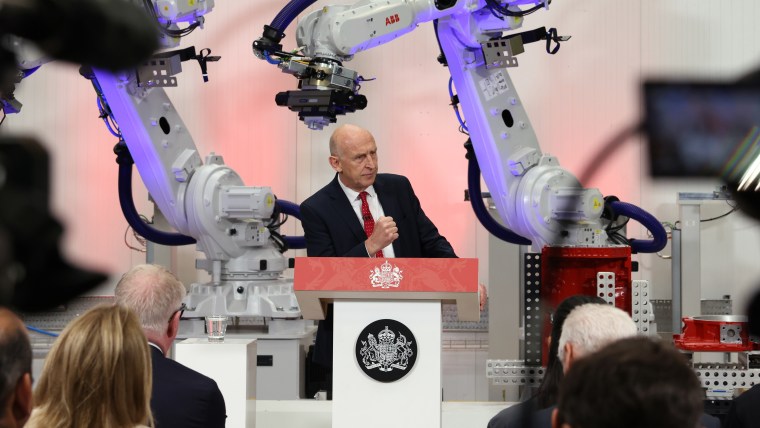 A bald man in a suit speaking at a podium with the Royal Coat of Arms, flanked by two ABB industrial robots.