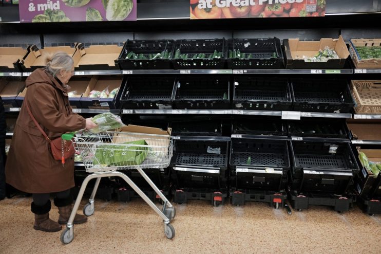 Shopper places produce into a cart in a grocery store aisle with mostly empty shelves, indicating supply shortages.