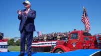 Donald Trump on a blue stage, clapping, with uniformed first responders and a red FDNY fire truck.