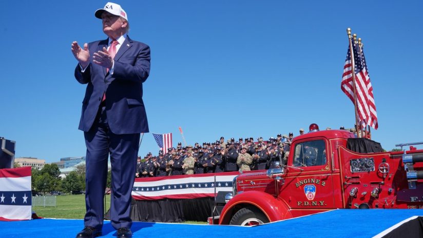 Donald Trump on a blue stage, clapping, with uniformed first responders and a red FDNY fire truck.