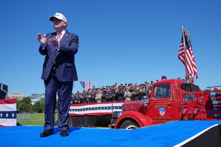 Donald Trump on a blue stage, clapping, with uniformed first responders and a red FDNY fire truck.