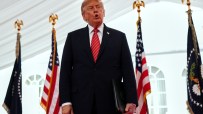 Donald Trump speaking at an event with US and presidential flags, holding a black folder
