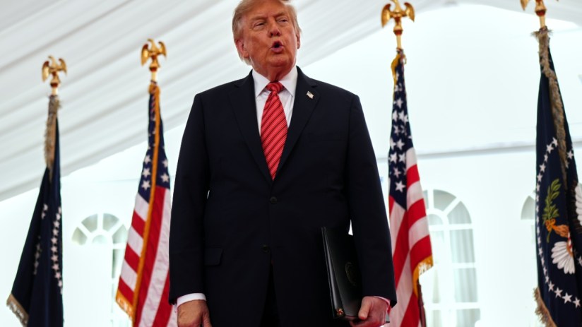 Donald Trump speaking at an event with US and presidential flags, holding a black folder
