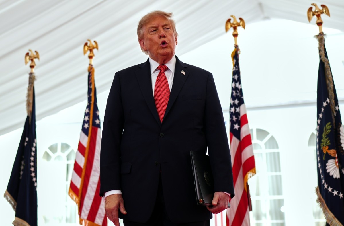 Donald Trump speaking at an event with US and presidential flags, holding a black folder