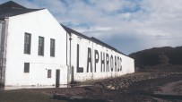 White Laphroaig distillery building with large black lettering on its side, beside a rocky shoreline and hills.