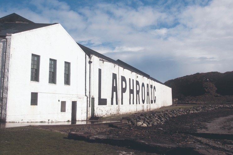 White Laphroaig distillery building with large black lettering on its side, beside a rocky shoreline and hills.