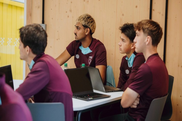 Young men in West Ham United shirts watching a presentation with laptops on a table in a modern room.