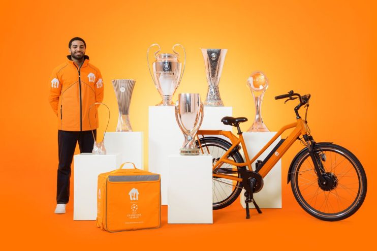 Delivery driver in orange jacket with UEFA Champions League trophies, delivery bag, and orange bicycle.