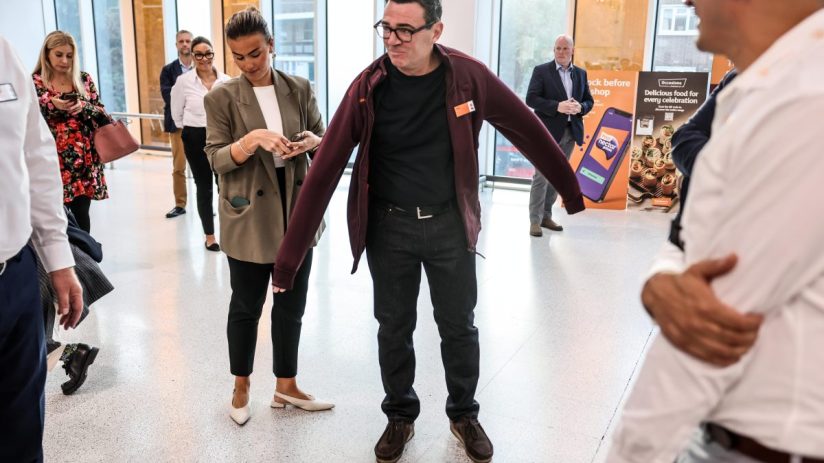 Andy Burnham in a Sainsburys fleece, adjusting his jacket, at a retail event with other attendees.