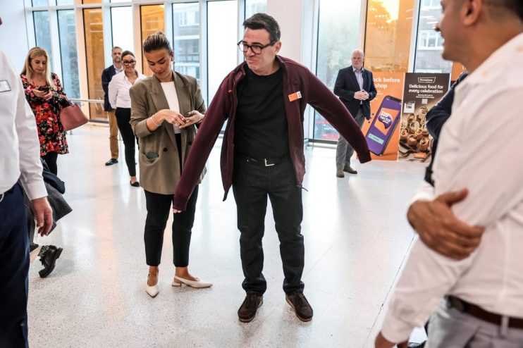Andy Burnham in a Sainsburys fleece, adjusting his jacket, at a retail event with other attendees.