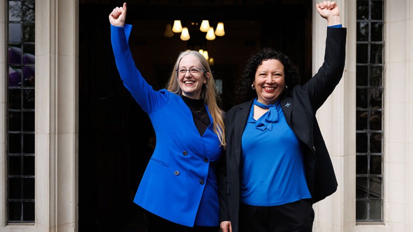 Two women, one with long blonde hair and glasses, the other with dark curly hair, raise their arms in triumph.