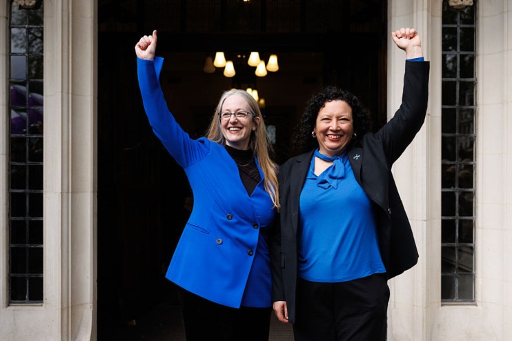 Two women, one with long blonde hair and glasses, the other with dark curly hair, raise their arms in triumph.