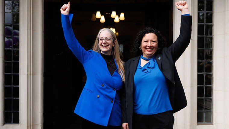 Two women, one with long blonde hair and glasses, the other with dark curly hair, raise their arms in triumph.