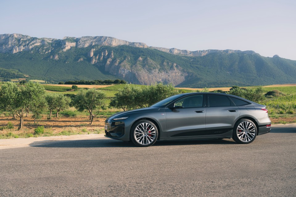 Grey Audi A6 Sportback e-tron parked on a road with mountains, olive groves, and vineyards in the background.