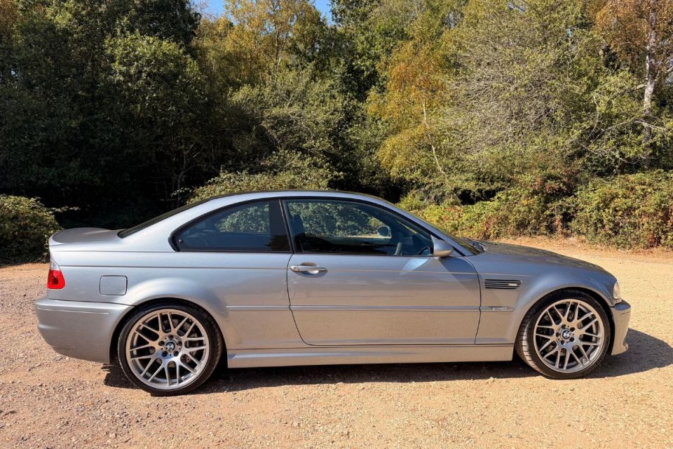 Silver BMW CSL1 coupe parked on a dirt road with green and autumn trees in the background.