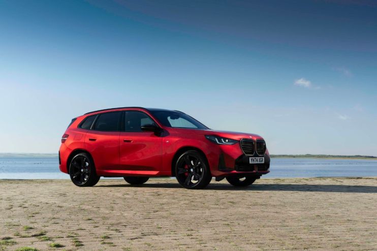 Red BMW X3 SUV parked on a paved surface next to calm water under a clear blue sky