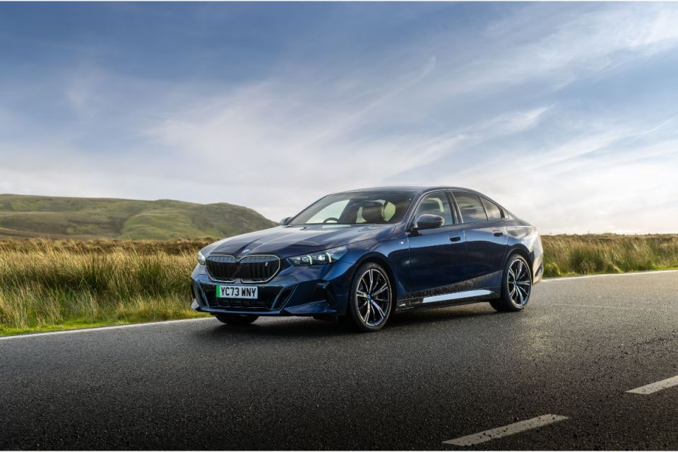 Blue BMW i5 electric sedan on an open road with green hills and a blue sky in the background.