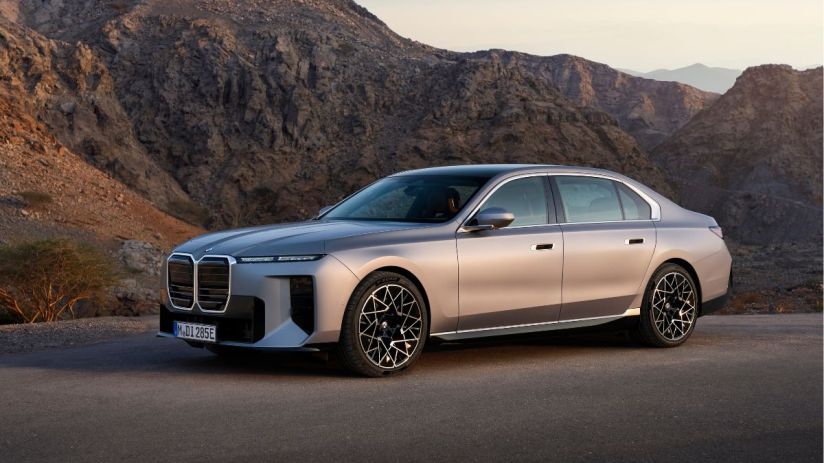 A sleek, silver BMW i7 luxury sedan parked on an asphalt road with rugged mountains in the background.