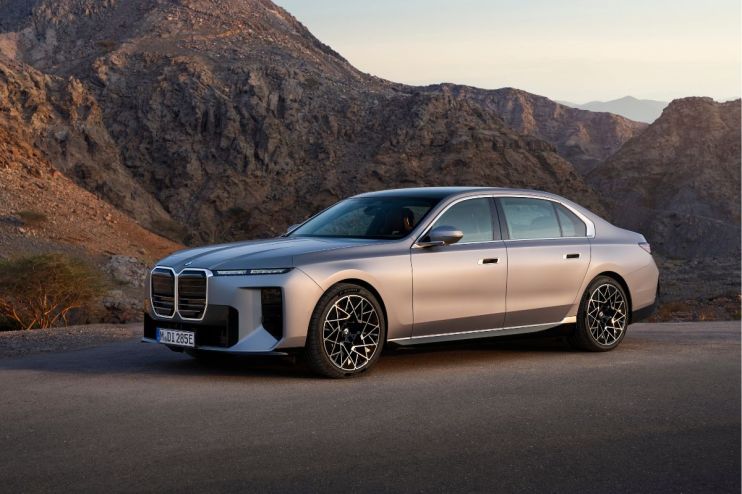A sleek, silver BMW i7 luxury sedan parked on an asphalt road with rugged mountains in the background.