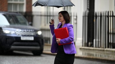 Bridget Phillipson, in a purple jacket, holds a red folder and black umbrella in the rain.
