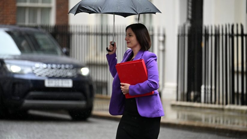 Bridget Phillipson, in a purple jacket, holds a red folder and black umbrella in the rain.