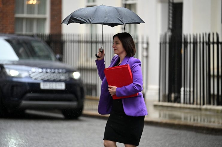 Bridget Phillipson, in a purple jacket, holds a red folder and black umbrella in the rain.