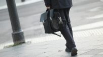 Man in dark suit carrying a black leather briefcase, walking on a city sidewalk.