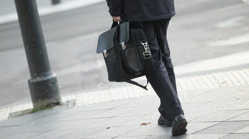 Man in dark suit carrying a black leather briefcase, walking on a city sidewalk.
