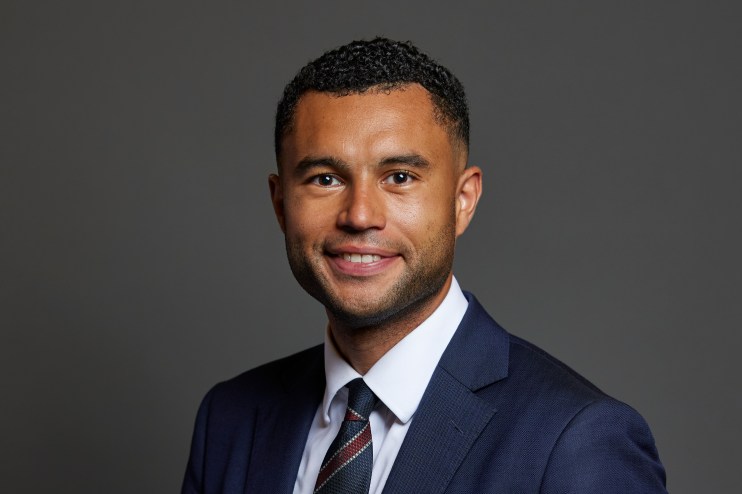 Callum Anderson, a smiling man in a navy suit and patterned tie, against a dark gray background.