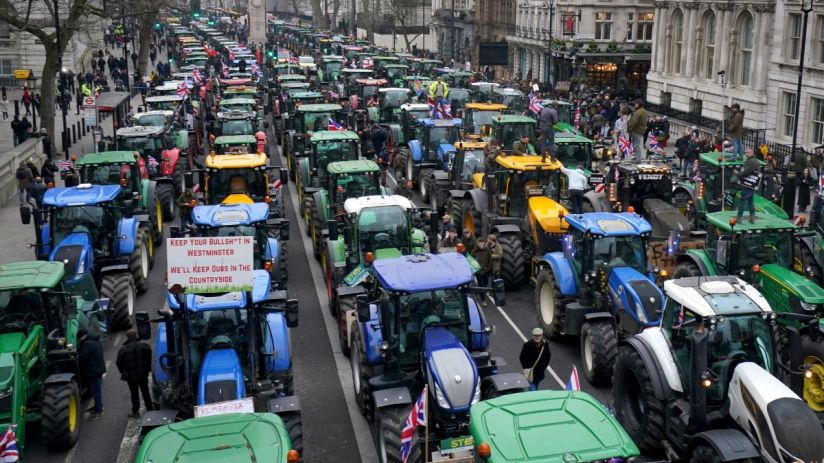Long line of tractors, many with Union Jack flags, protesting in a city street, one sign reads Keep your bullsh*t in Westm...