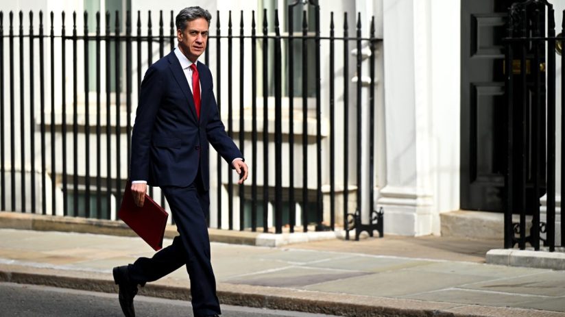 Ed Miliband, in a suit and red tie, strides past Downing Street railings with a red folder in hand.