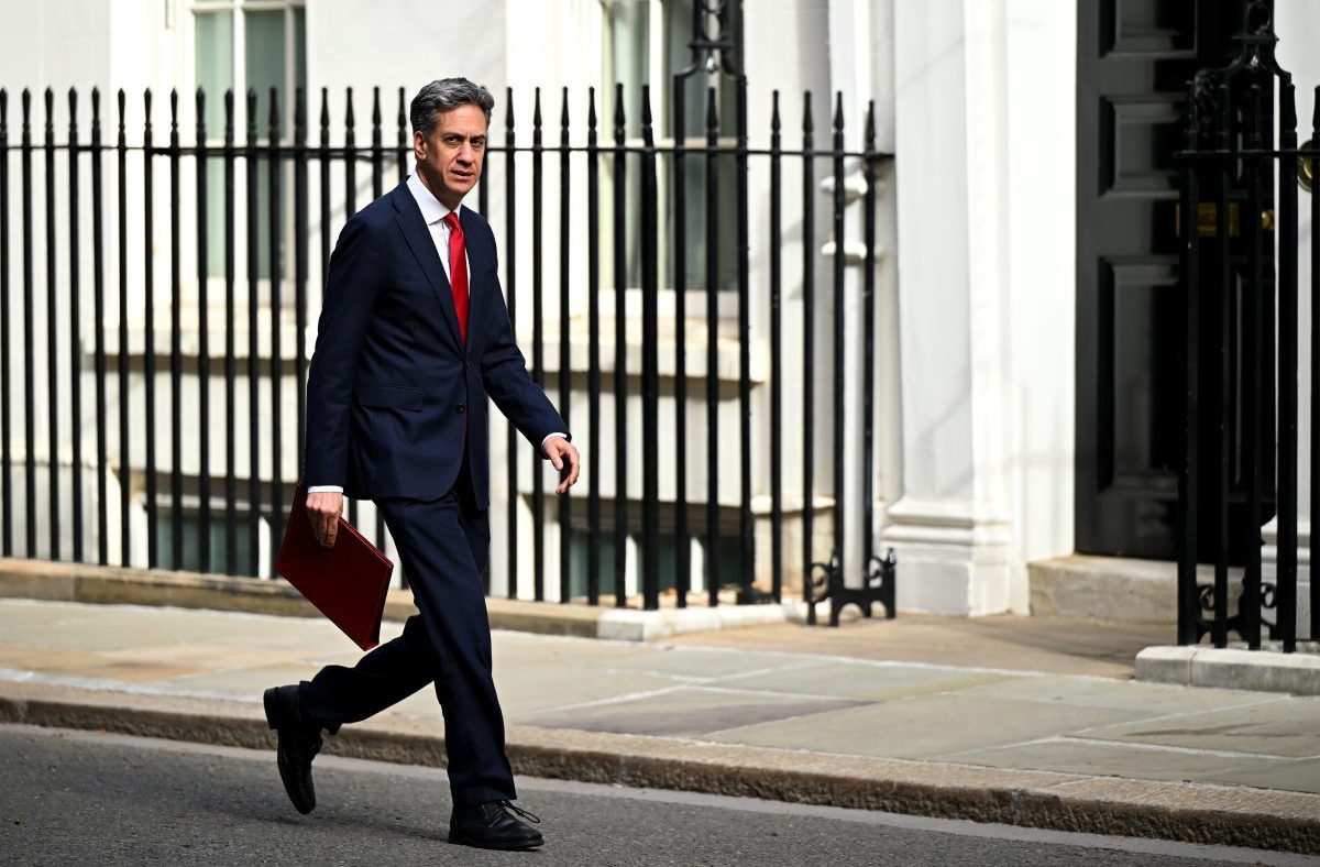 Ed Miliband, in a suit and red tie, strides past Downing Street railings with a red folder in hand.