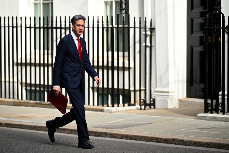 Ed Miliband in a suit and red tie, carrying a red folder, walking past railings outside Downing Street